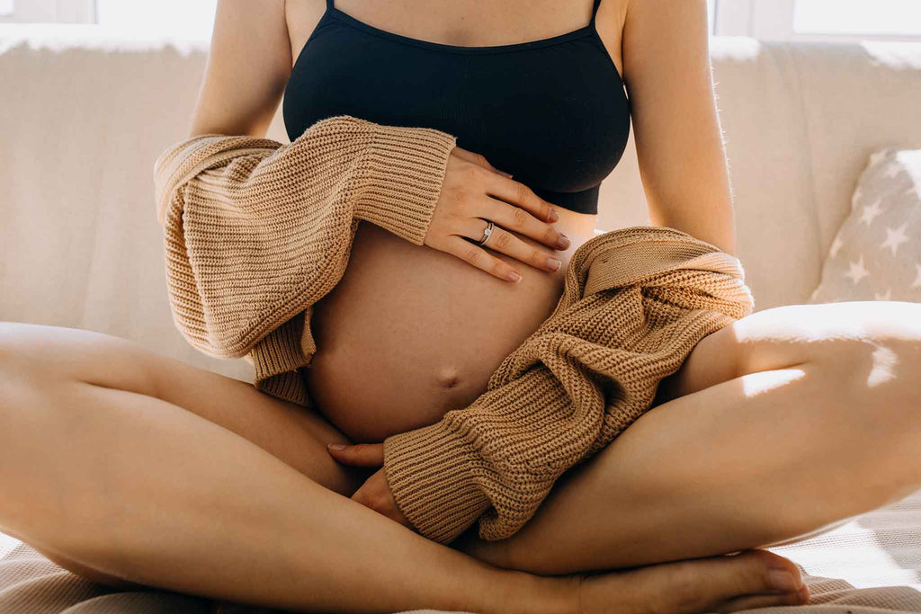 Eine schwangere Frau sitzt mit gekreuzten Beinen auf einem Sofa und streichelt sanft ihren Bauch.