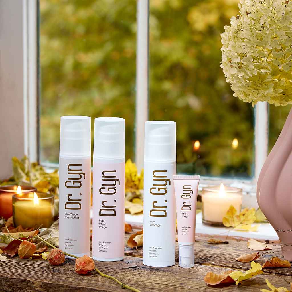 „Dr. Gyn“ Körperpflegeprodukte gegen trockene Haut vor einem Fenster mit Herbstblättern.