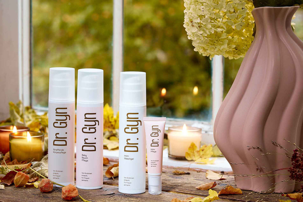 „Dr. Gyn“ Körperpflegeprodukte gegen trockene Haut vor einem Fenster mit Herbstblättern.