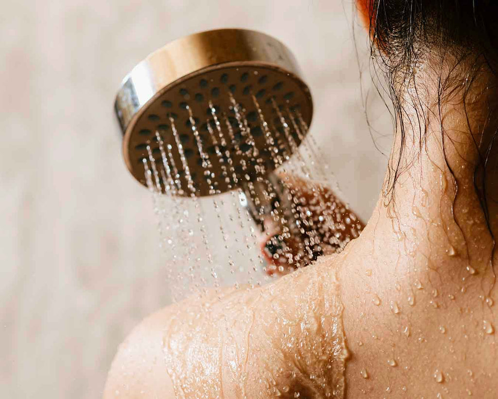Frau, die unter der Dusche steht, wobei Wasser aus einer Handbrause auf ihre Schultern herabfließt. 